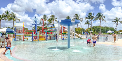 A vibrant water park scene with slides, fountains, and palm trees under a sunny sky, filled with families enjoying the water.