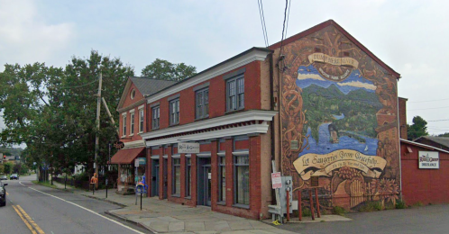 A brick building with a colorful mural, located on a street with trees and other shops nearby.