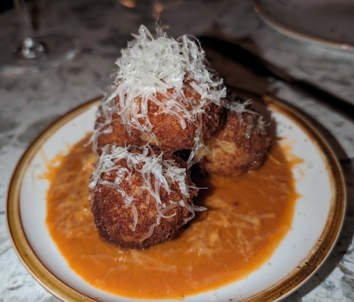Four golden-brown croquettes topped with shredded cheese, served on a plate with a rich orange sauce.