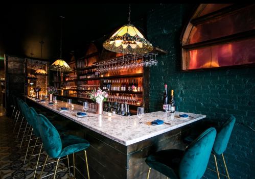 A stylish bar with a marble countertop, green velvet chairs, and vintage lighting, featuring shelves of bottles in the background.