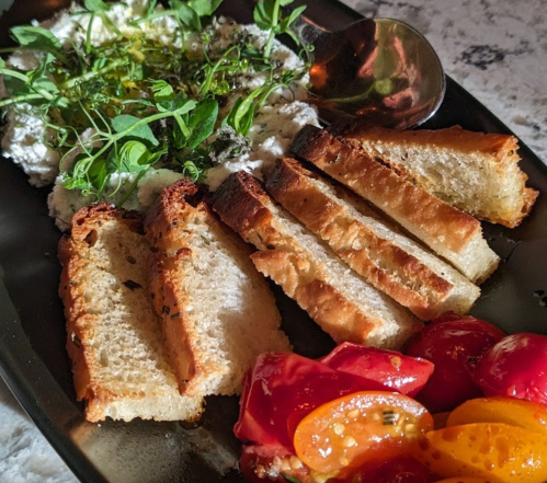 A plate featuring sliced toasted bread, creamy spread, fresh herbs, and colorful tomatoes.