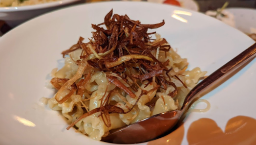 A bowl of pasta topped with crispy fried onions, served with a spoon.