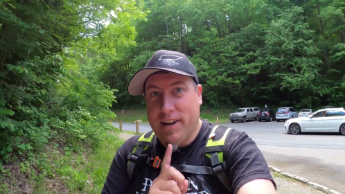 A person in a cap and backpack gestures with a finger to their lips, standing in a green, wooded area near parked cars.