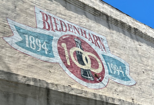 Faded mural featuring "Biedenharn" with dates 1894 and 1994, and a bottle icon, against a blue sky.