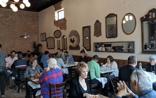 A bustling restaurant interior with diners at tables, exposed brick walls, and vintage mirrors.