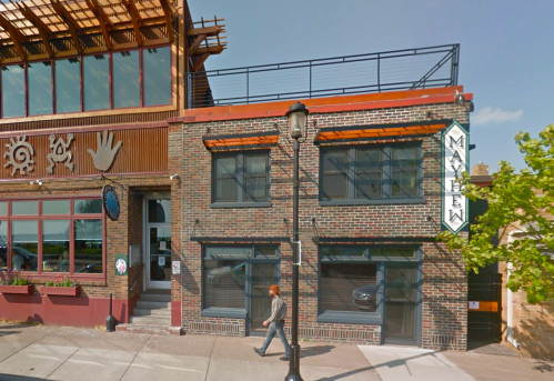A brick building with large windows and a sign reading "Mayhew," next to a modern structure with a rooftop terrace.