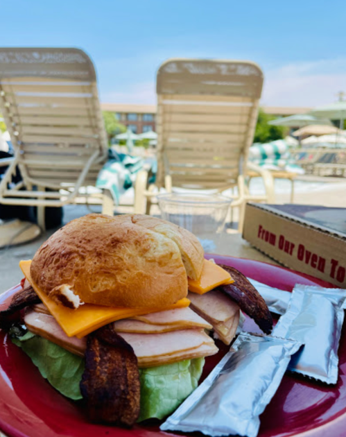 A close-up of a sandwich with turkey, cheese, and lettuce on a red plate, with poolside lounge chairs in the background.