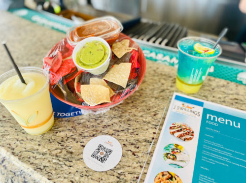 A table with a basket of chips and dips, two colorful drinks, and a menu on a granite surface.