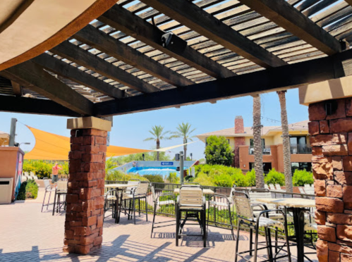 A shaded outdoor seating area with tables, palm trees, and a pool in the background on a sunny day.