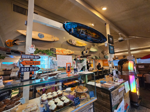 A cozy café interior with surfboards on the wall, a display of baked goods, and a colorful jukebox.