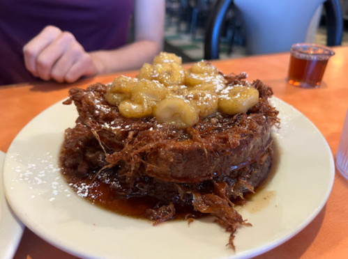 A plate of bread pudding topped with caramelized bananas and drizzled with syrup, served in a casual dining setting.
