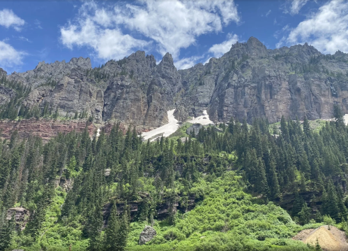 A rugged mountain landscape with steep cliffs, patches of snow, and lush green trees in the foreground.