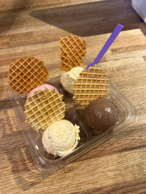 A clear container holds three scoops of ice cream topped with waffle cones on a wooden surface.