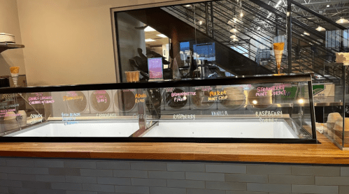 Ice cream shop display with empty tubs and colorful flavor labels on a wooden counter. Modern interior in the background.