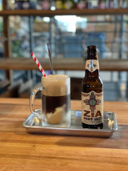 A frosty mug of root beer with foam, a striped straw, and a bottle of Zia Root Beer on a metal tray.