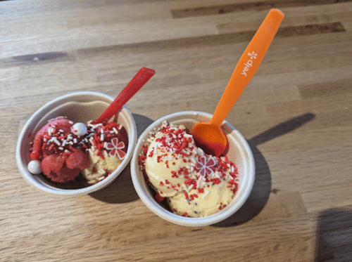 Two small cups of ice cream: one red with sprinkles and one yellow with red sprinkles, both with plastic spoons.