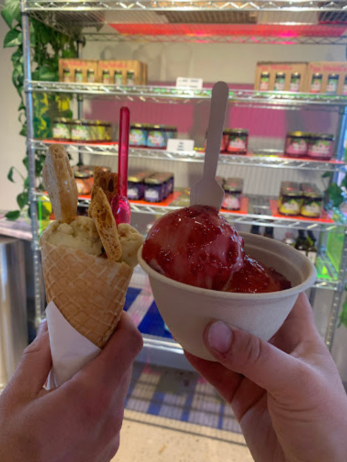 Two hands hold ice cream: a cone with toppings and a bowl of strawberry ice cream, with colorful jars in the background.