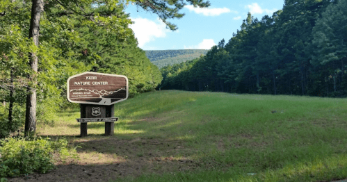 Sign for Kerr Nature Center at the edge of a grassy area, surrounded by tall trees and mountains in the background.