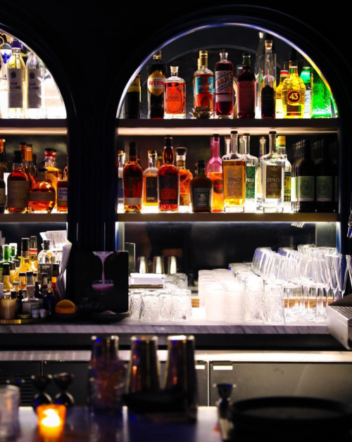A well-stocked bar with various bottles of liquor on shelves, illuminated by soft lighting, and glassware in the foreground.