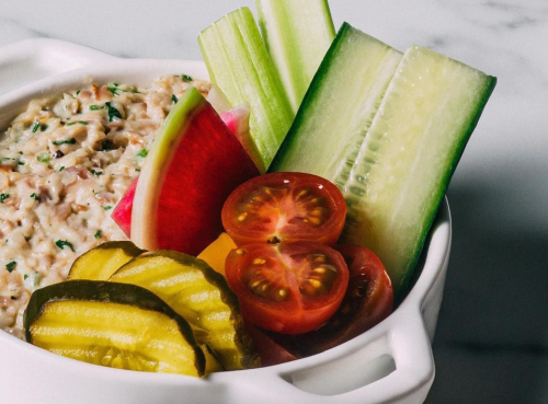 A bowl of creamy dip surrounded by sliced cucumbers, cherry tomatoes, pickles, and watermelon radish.