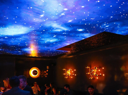 A dimly lit restaurant with a starry ceiling and stylish wall lights, featuring patrons enjoying their evening.