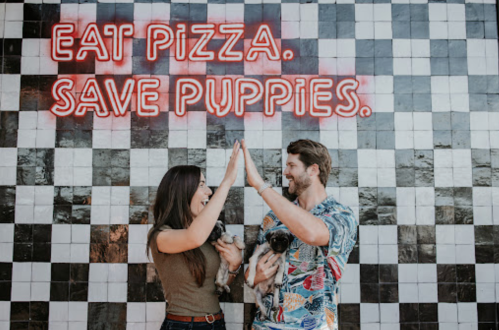 A couple high-fives while holding puppies in front of a wall with a neon sign that says "Eat Pizza. Save Puppies."