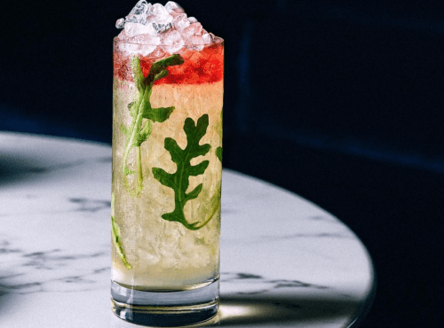 A tall cocktail glass filled with a layered drink, ice, and garnished with green herbs on a marble table.
