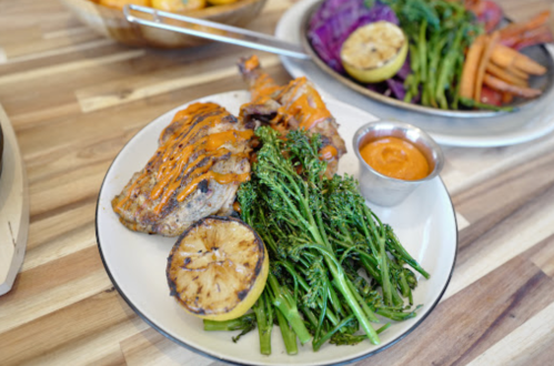 Grilled chicken with sauce, broccolini, and charred lemon on a plate, served with a small cup of dipping sauce.