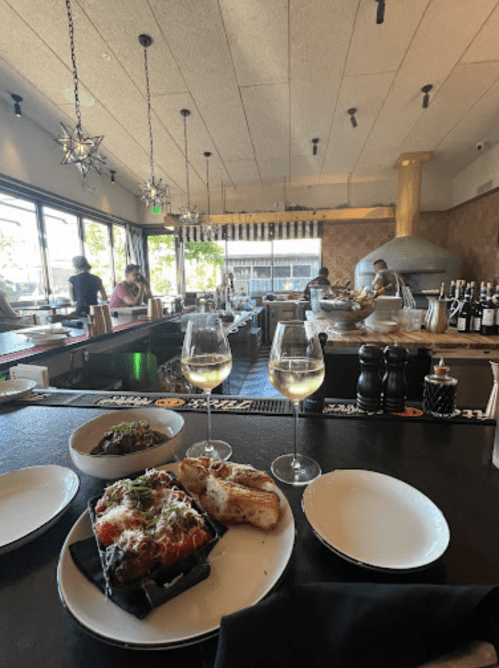 A restaurant interior with two glasses of white wine, a plate of food, and a view of diners and a pizza oven.