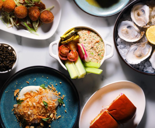 A spread of gourmet dishes including oysters, a seafood dip, fried snacks, and a sandwich on a marble table.