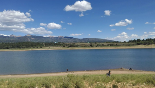 A serene lake surrounded by mountains and trees, with people enjoying the shoreline under a blue sky with clouds.