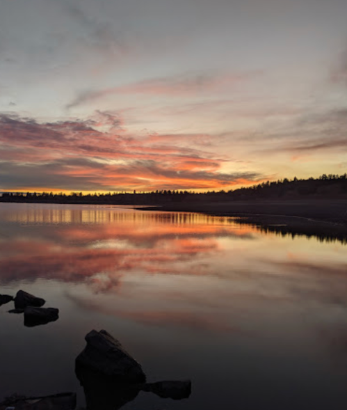 A serene sunset over a calm lake, with colorful clouds reflecting on the water's surface and rocky shoreline.