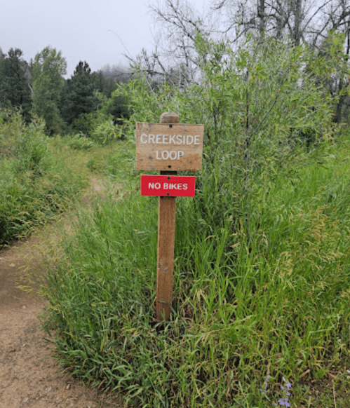 A wooden sign marking the Creekside Loop trail, with a "No Bikes" notice, surrounded by tall green grass and trees.