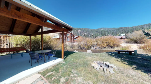 A shaded patio with a table and chairs overlooks a stream and fire pit, surrounded by trees and mountains.