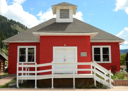 A red, one-story building with a white door and windows, featuring a small cupola on the roof and a ramp in front.