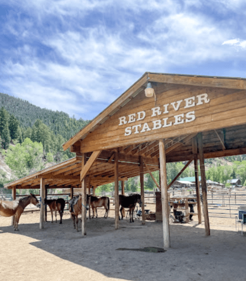 A wooden stable with the sign "Red River Stables," surrounded by horses and a scenic mountain backdrop.