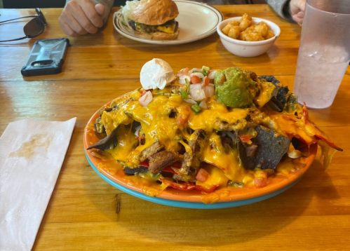 A colorful plate of nachos topped with cheese, guacamole, sour cream, and diced tomatoes, with a burger and fries in the background.