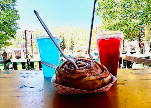 A cinnamon roll with two forks, flanked by blue and red drinks, set against a scenic outdoor backdrop.