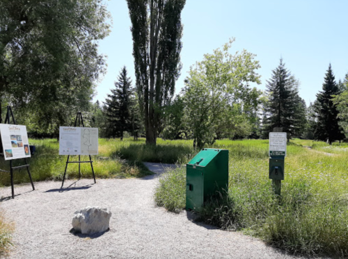 A green recycling bin and informational signs in a grassy park area with trees and a clear blue sky.