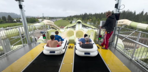 Two people sit in inflatable rafts at the top of a water slide, while an attendant prepares to launch them.