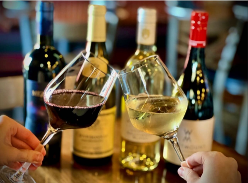 Two glasses of wine clink together in front of several wine bottles on a table.