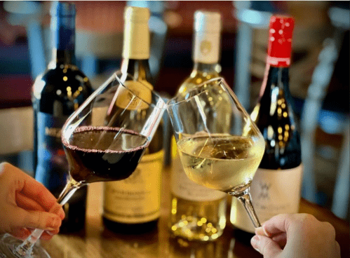 Two glasses of wine clink together in front of several wine bottles on a table.