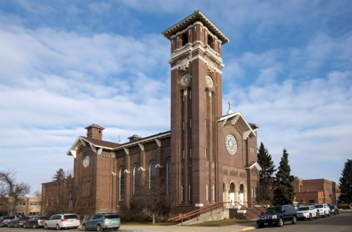 Historic brick church with a tall clock tower, surrounded by trees and cars under a partly cloudy sky.