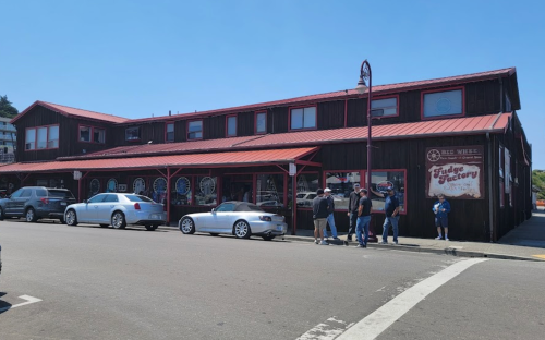 A wooden building with a red roof, featuring shops and people standing outside on a sunny day.