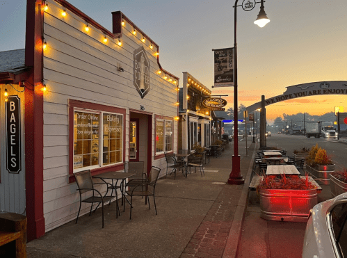A quaint street scene at dawn featuring a bagel shop with outdoor seating and warm string lights.