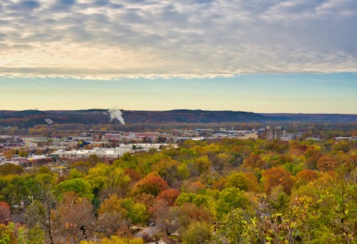 A panoramic view of a colorful autumn landscape with a town nestled among trees and rolling hills under a cloudy sky.