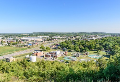 Aerial view of a cityscape with industrial buildings, green spaces, and water treatment facilities under a clear blue sky.