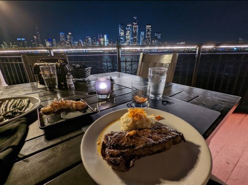 A plate with steak and mashed potatoes on a table, with a city skyline view at night in the background.
