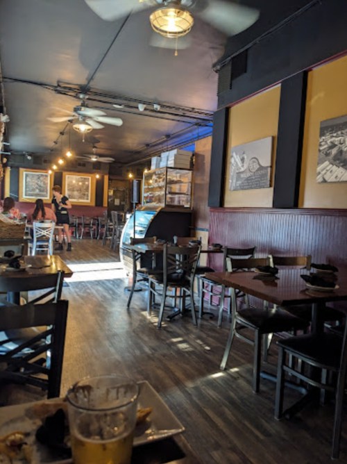 Interior of a restaurant with wooden tables, chairs, and a display case, featuring warm lighting and a casual atmosphere.