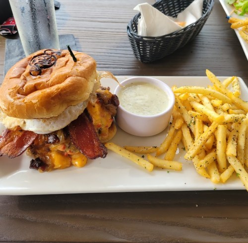 A hearty burger with bacon, cheese, and an egg, served with a side of fries and dipping sauce on a white plate.